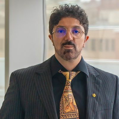 Man in black jacket, colorful yellow tie, wearing glasses, has a beard.