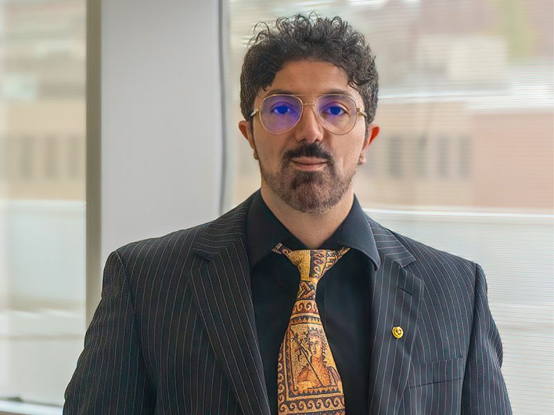 Man in black jacket, colorful yellow tie, wearing glasses, has a beard.