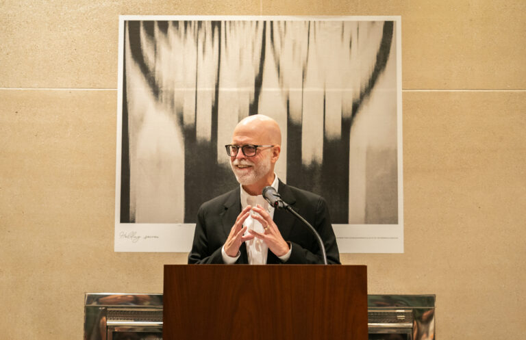 Person stands at a podium with a microphone, speaking in front of a large abstract black-and-white photograph mounted on a beige wall.