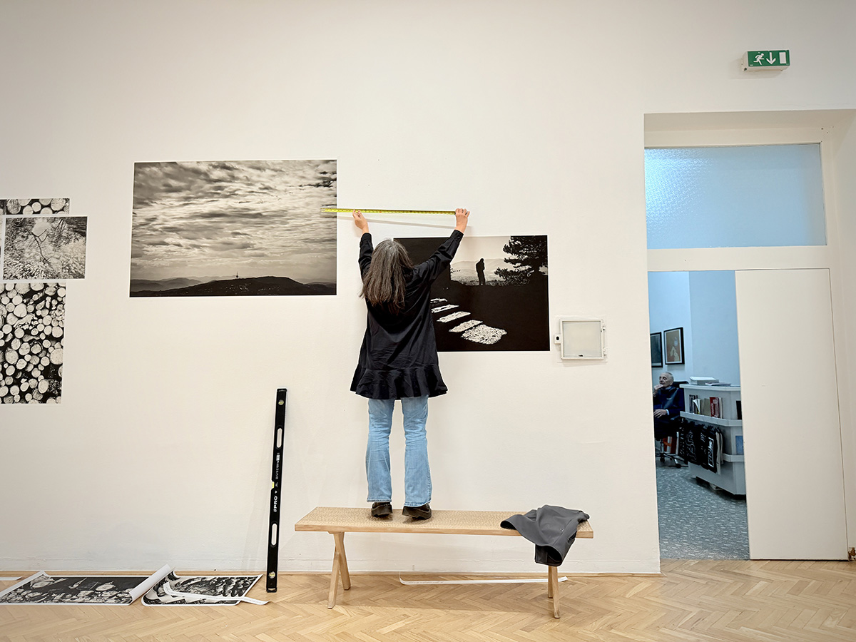 Person stands on a wooden bench using a measuring tape to position large black-and-white photographs on a gallery wall, with tools and prints scattered nearby.