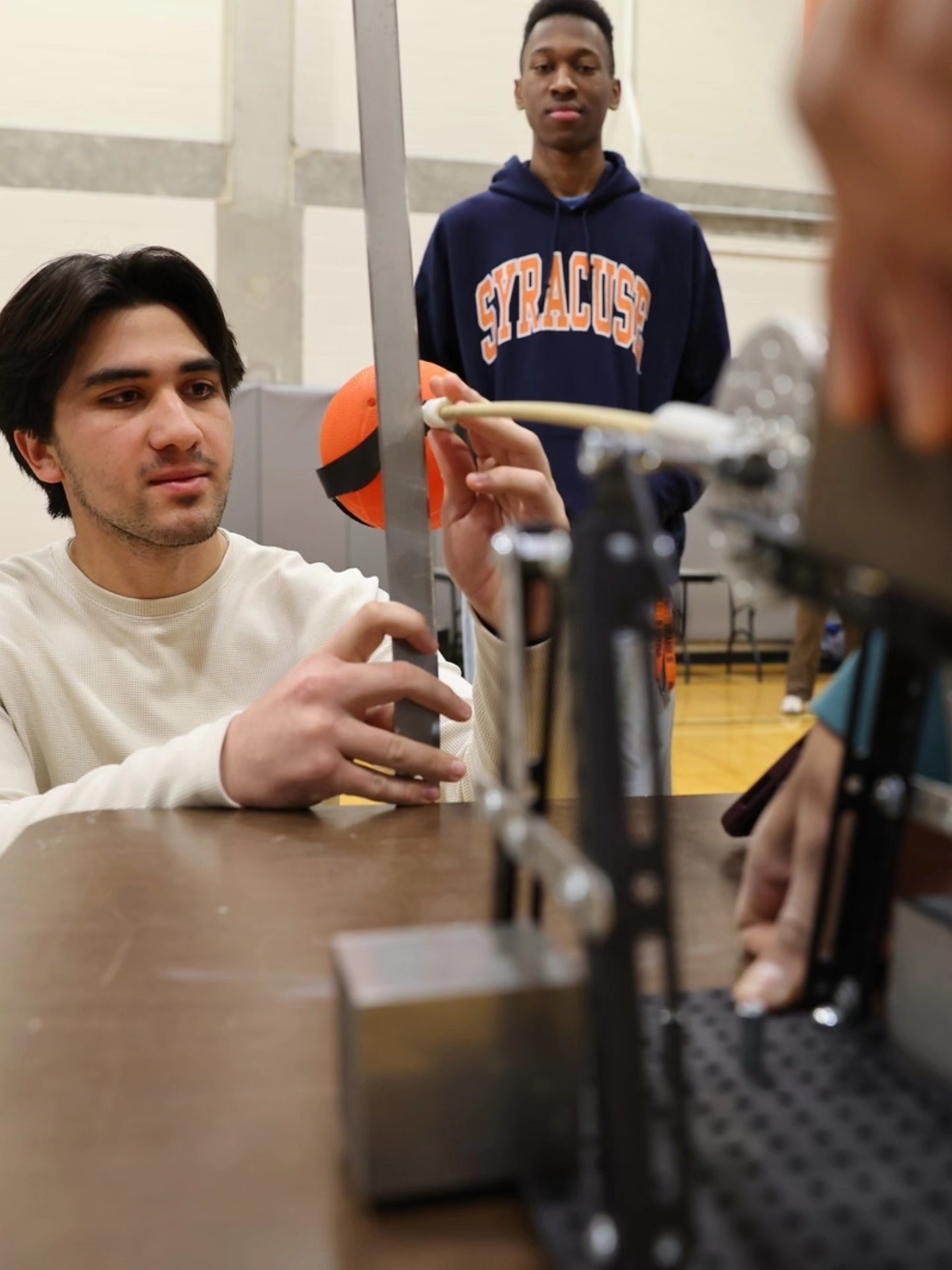 Close-up of a student adjusting a metal frame on a tabletop basketball launcher while another person wearing a Syracuse hoodie stands in the background.