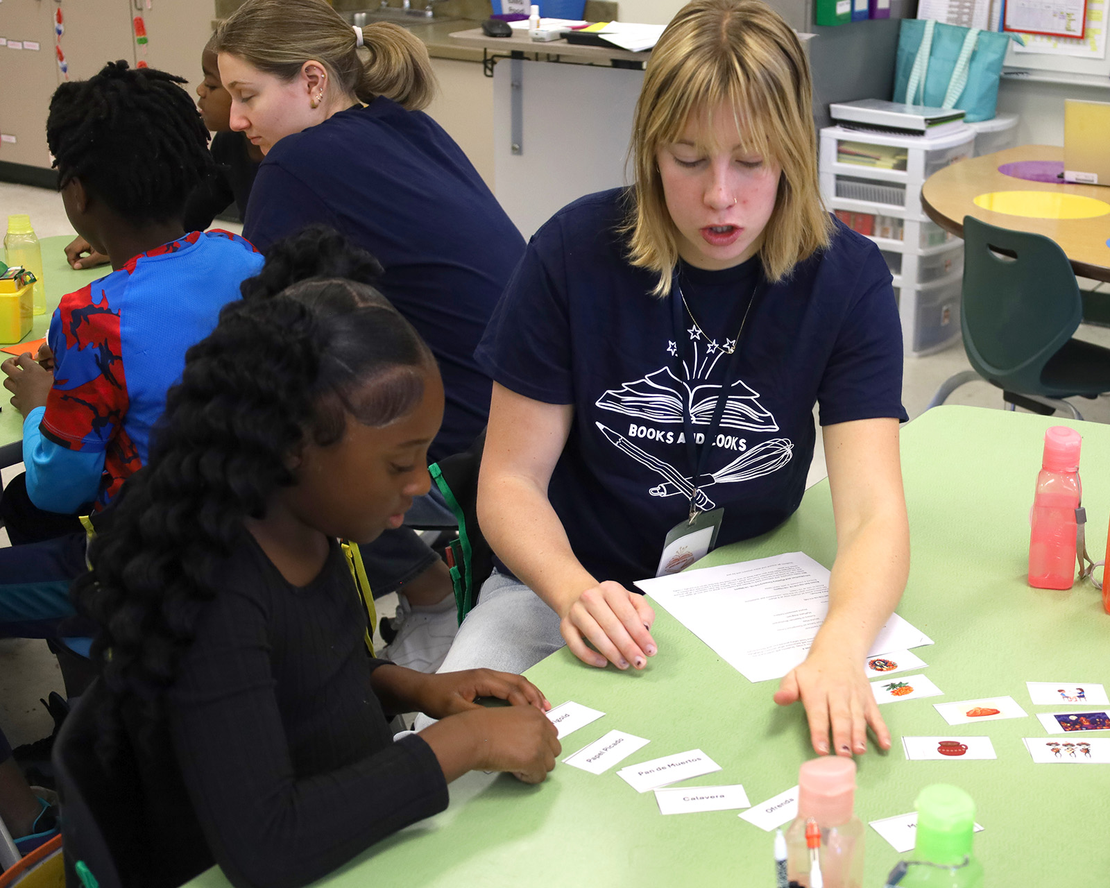 An adult and a child sit at a classroom table working on a nutrition activity with word cards and food images. Other students are seated at nearby tables with water bottles and papers.