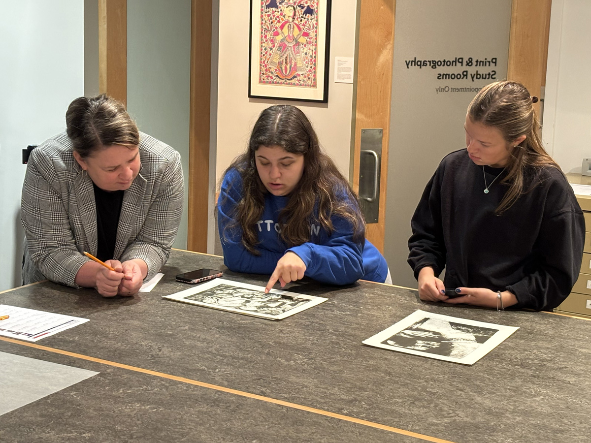 Three people standing at a table in an art study room, closely examining black-and-white prints and taking notes