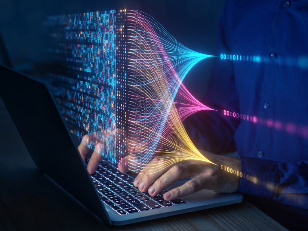 Hands typing on laptop with holographic visualization of colorful data streams, binary code, and flowing network lines representing artificial intelligence and machine learning processes.