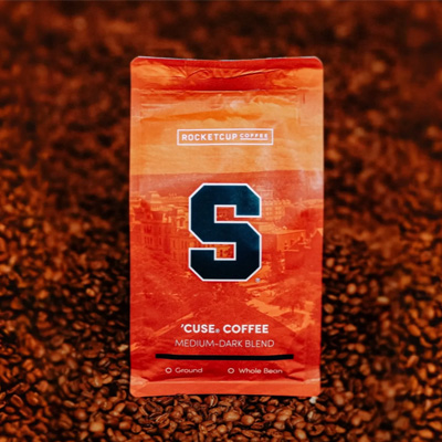 Orange coffee bag labeled Rocketcup Coffee with a large navy blue Block S and text reading ’Cuse Coffee, Medium-Dark Blend, placed on a background of roasted coffee beans