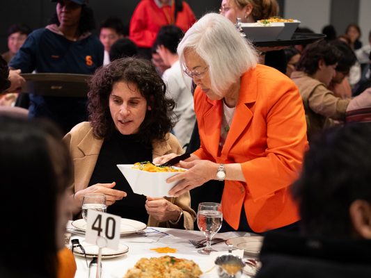 Two women, one standing and serving food to one seated at a table.