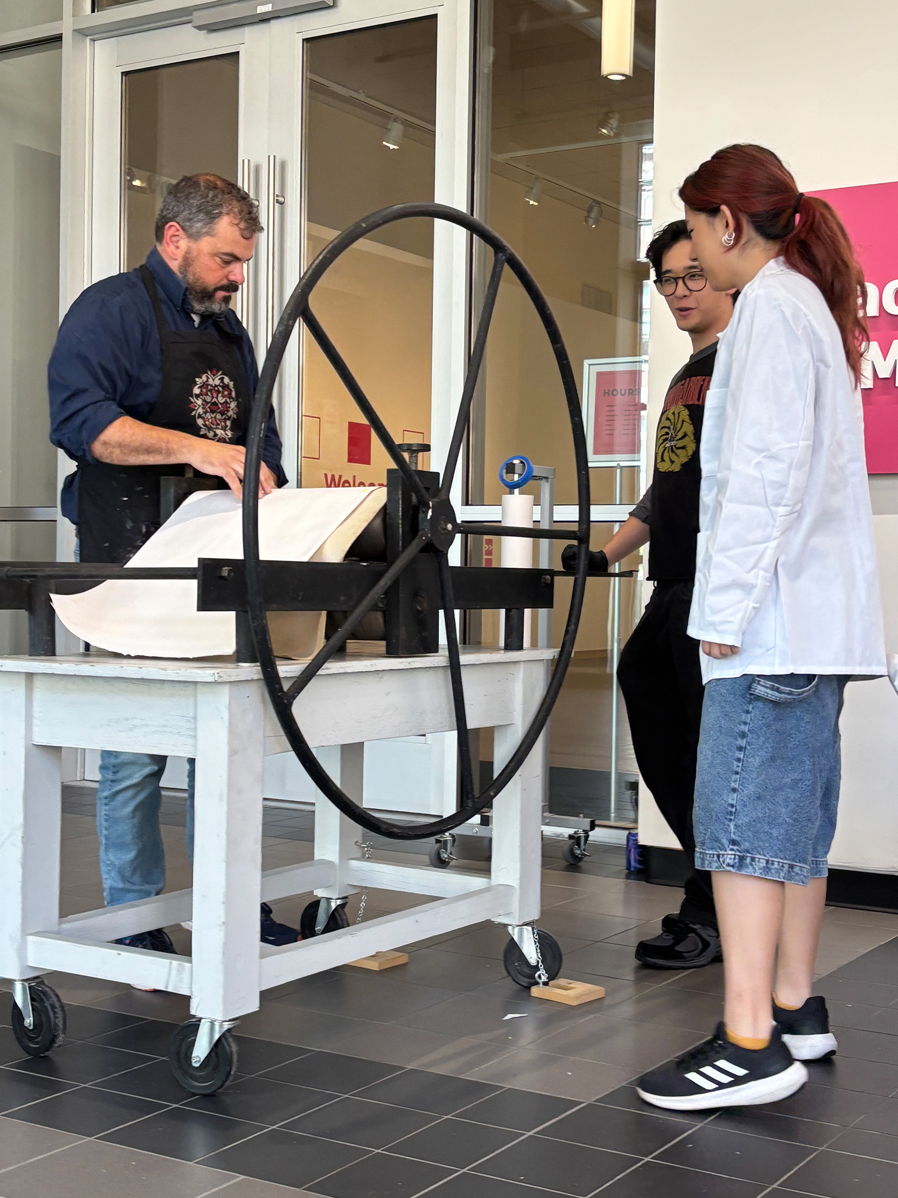 An instructor in a navy apron demonstrates a large wheel printing press to two students in a bright gallery space.