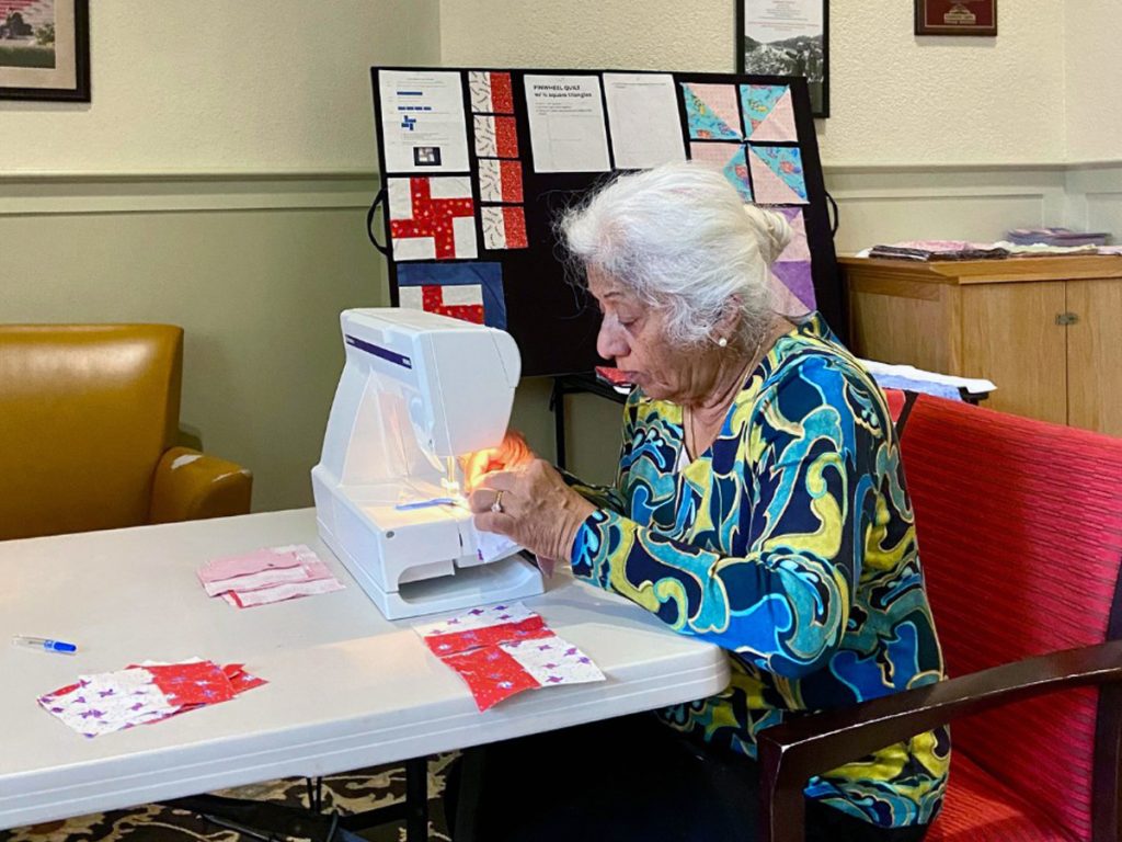 Woman in colorful patterned top works at sewing machine, crafting quilted fabric squares displayed on table and board behind her.