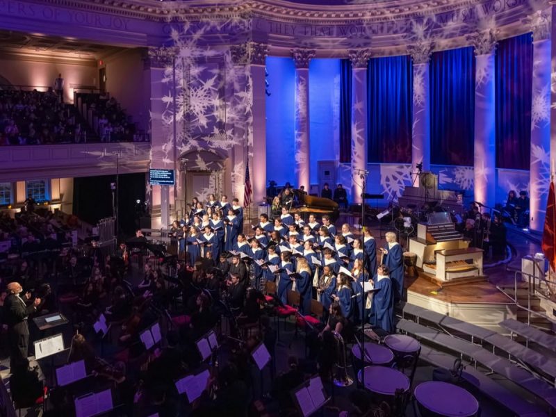 Choir in blue robes performing on stage with an orchestra in a grand hall decorated with snowflake light projections and blue curtains.