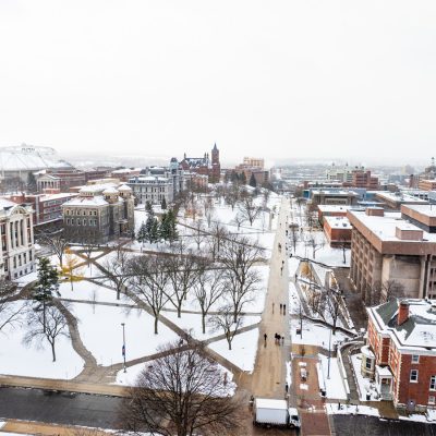 Aerial view of Syracuse University campus in winter, with snow-covered buildings, trees, and walkways along the main promenade