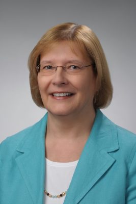 Person wearing a turquoise blazer over a white top with a beaded necklace, posed against a plain gray background.