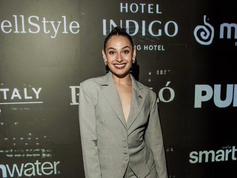 Person wearing a light gray suit with a deep neckline posing in front of a black backdrop featuring sponsor logos
