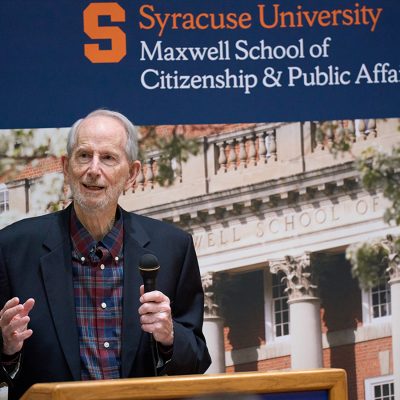 Man in dark coat speaks at a podium. A sign that says Syracuse University Maxwell School of Citizenship and Public Affairs is behind him.