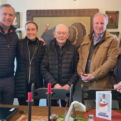 Five people standing closely together indoors in front of a wall with framed artwork featuring a large bird.