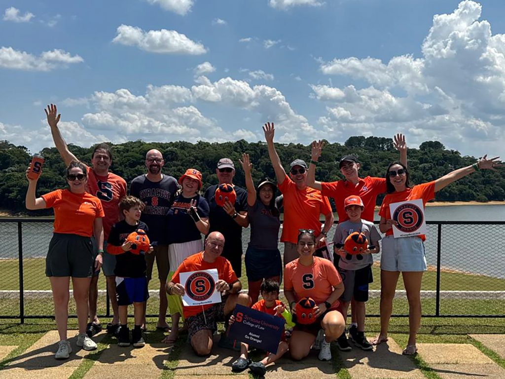 Large group of Syracuse University fans in orange and blue apparel pose together outdoors by lake, waving and holding Otto the Orange plush toys and Syracuse signs.