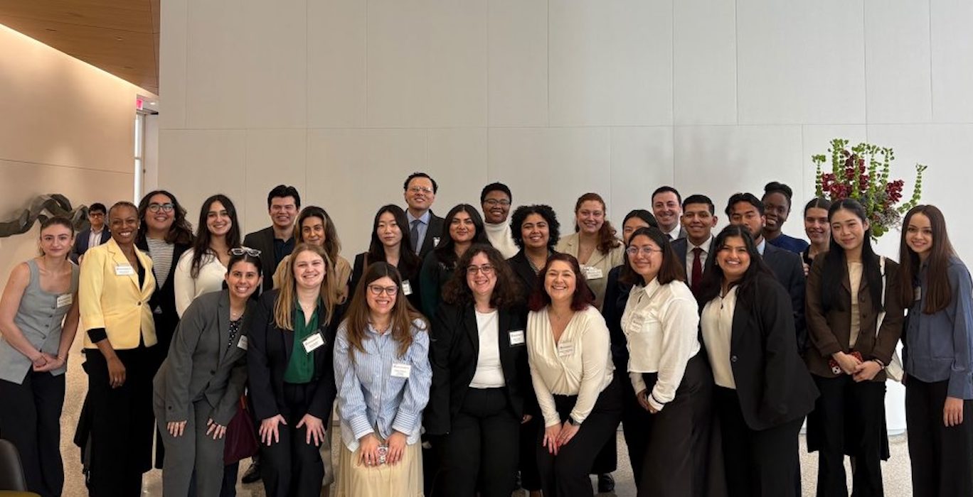 A large group of people dressed in professional attire posing together indoors against a plain wall. 