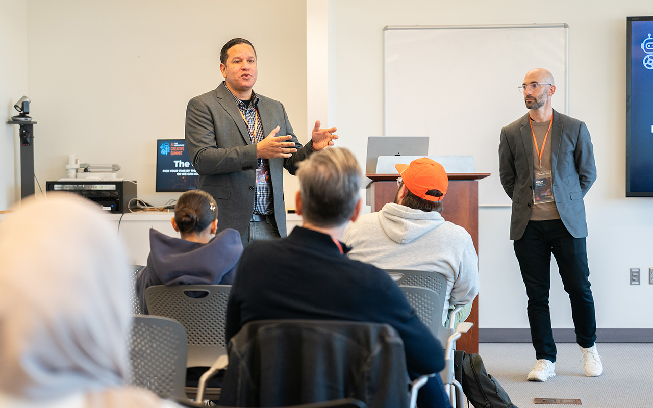 Speaker addressing an audience in a classroom-style setting with another presenter standing nearby. A podium, laptop, and whiteboard are visible at the front of the room