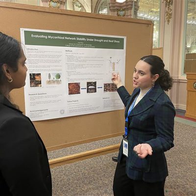 Student dressed in blue points to a poster mounter on a board as she makes a presentation to a woman dressed in black