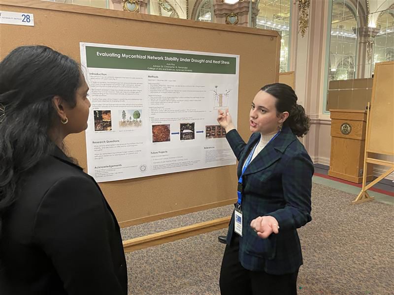 Student dressed in blue points to a poster mounter on a board as she makes a presentation to a woman dressed in black