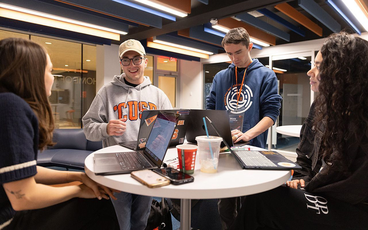 Group of students collaborating around a round table with laptops and drinks in a modern, brightly lit campus space