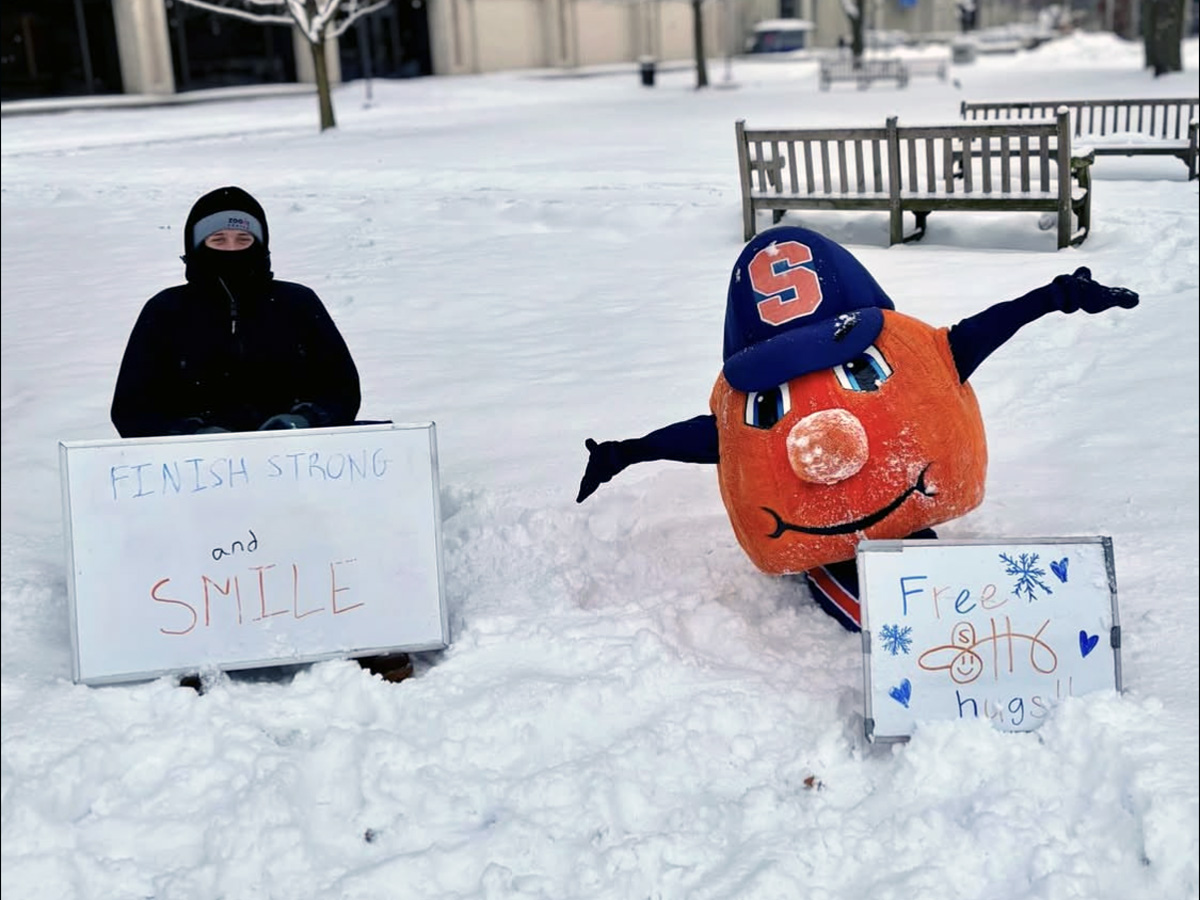 Otto the Orange mascot sits in the snow wearing a blue cap with an orange “S,” holding a sign that says “Free Otto hugs!” with snowflake and heart drawings. Next to Otto, another sign reads “Finish strong and smile.” Snow-covered benches and trees are visible in the background.