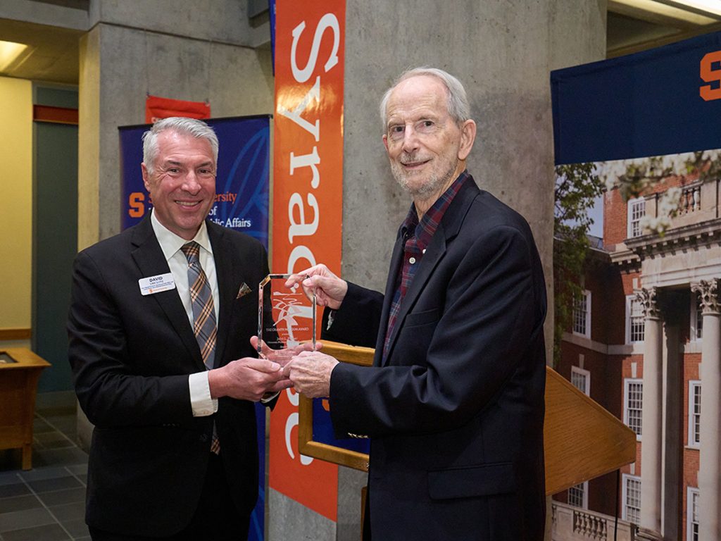 Man in dark jacket and smiling presents an award to a man in a dark jacket and smiling. The word Syracuse appears behind them.