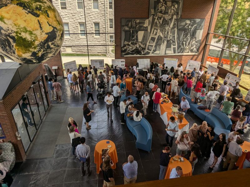 Crowded indoor event with people viewing research posters and networking around orange-covered tables in a bright atrium with large windows and artwork