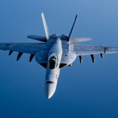 A U.S. military fighter jet flies directly toward the camera against a clear blue sky, showing its wings extended, cockpit centered, and twin vertical stabilizers at the rear.