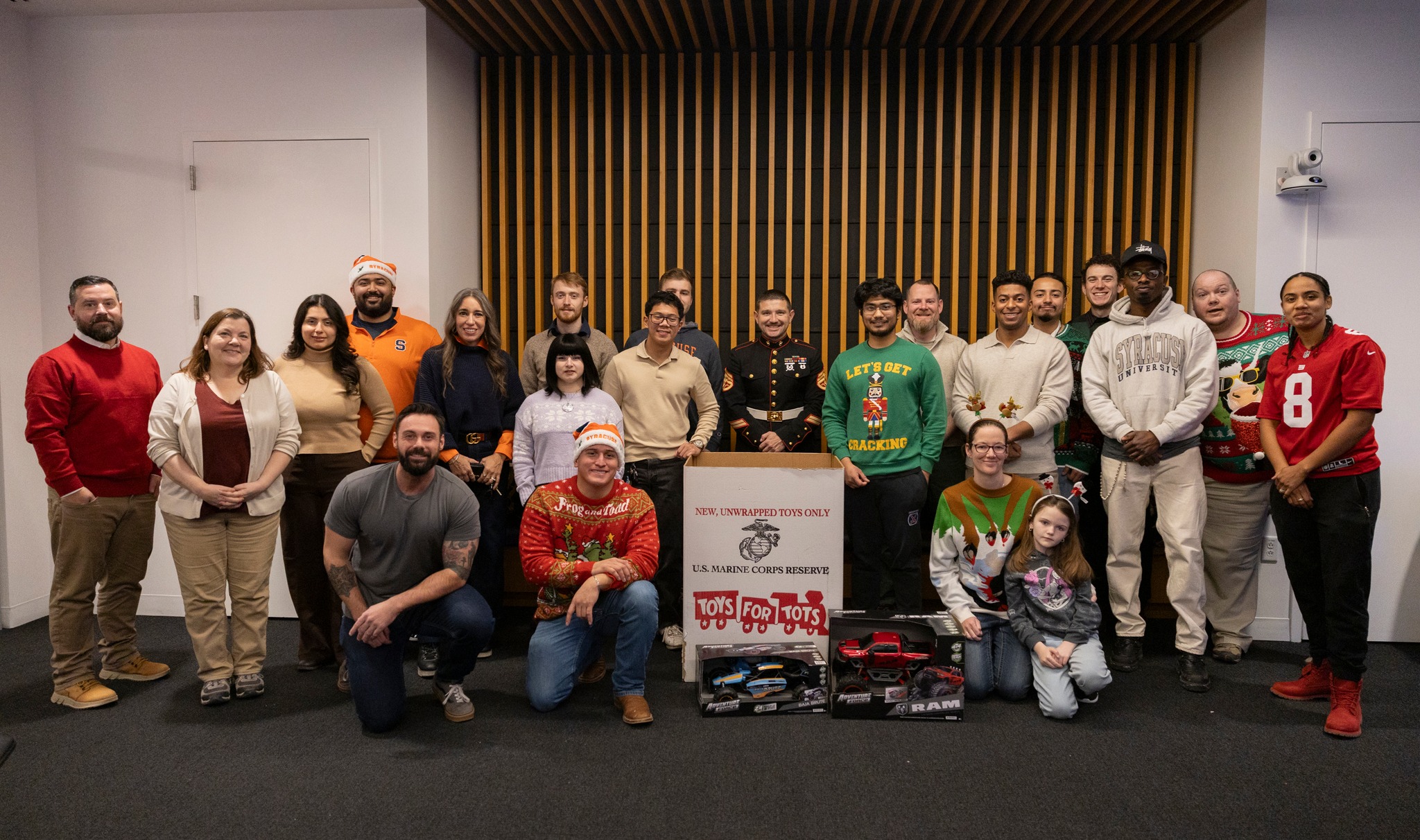 Group of people standing and kneeling around a Toys for Tots donation box with toy cars in front, inside a room with wood panel wall and gray carpet