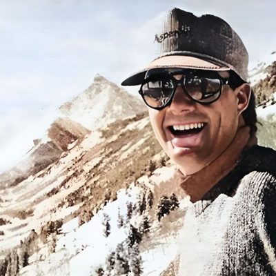 Man wearing baseball cap and sunglasses, smiling with mountains in the background.