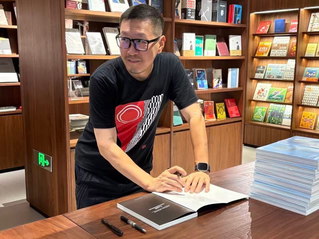 Man signing a book ata a table, pen in hand. There are books on the shelves behind him and a stack of books on the table.