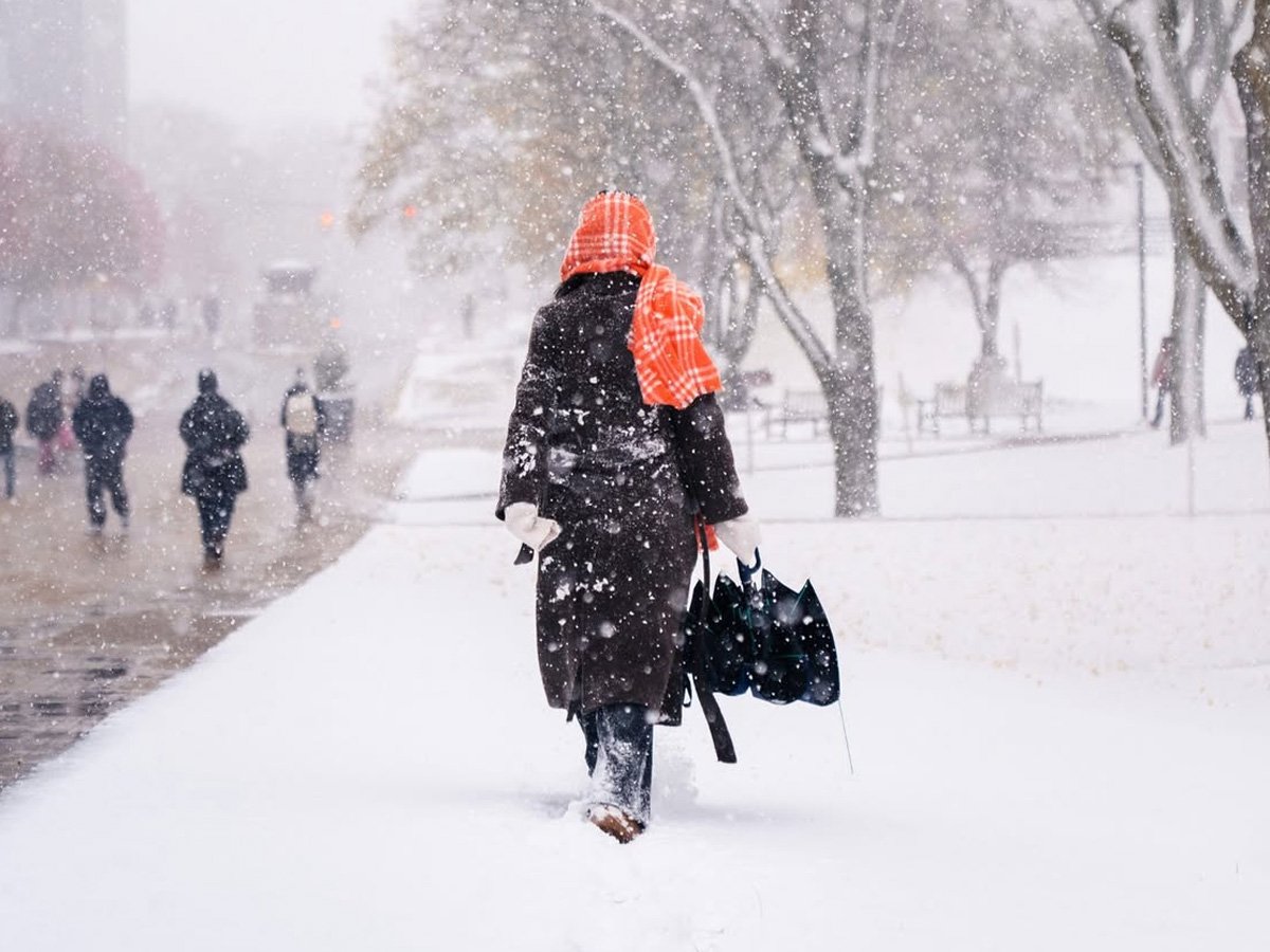 Person walking through heavy snowfall on a snow-covered sidewalk, wearing a dark coat and orange plaid scarf, carrying an umbrella