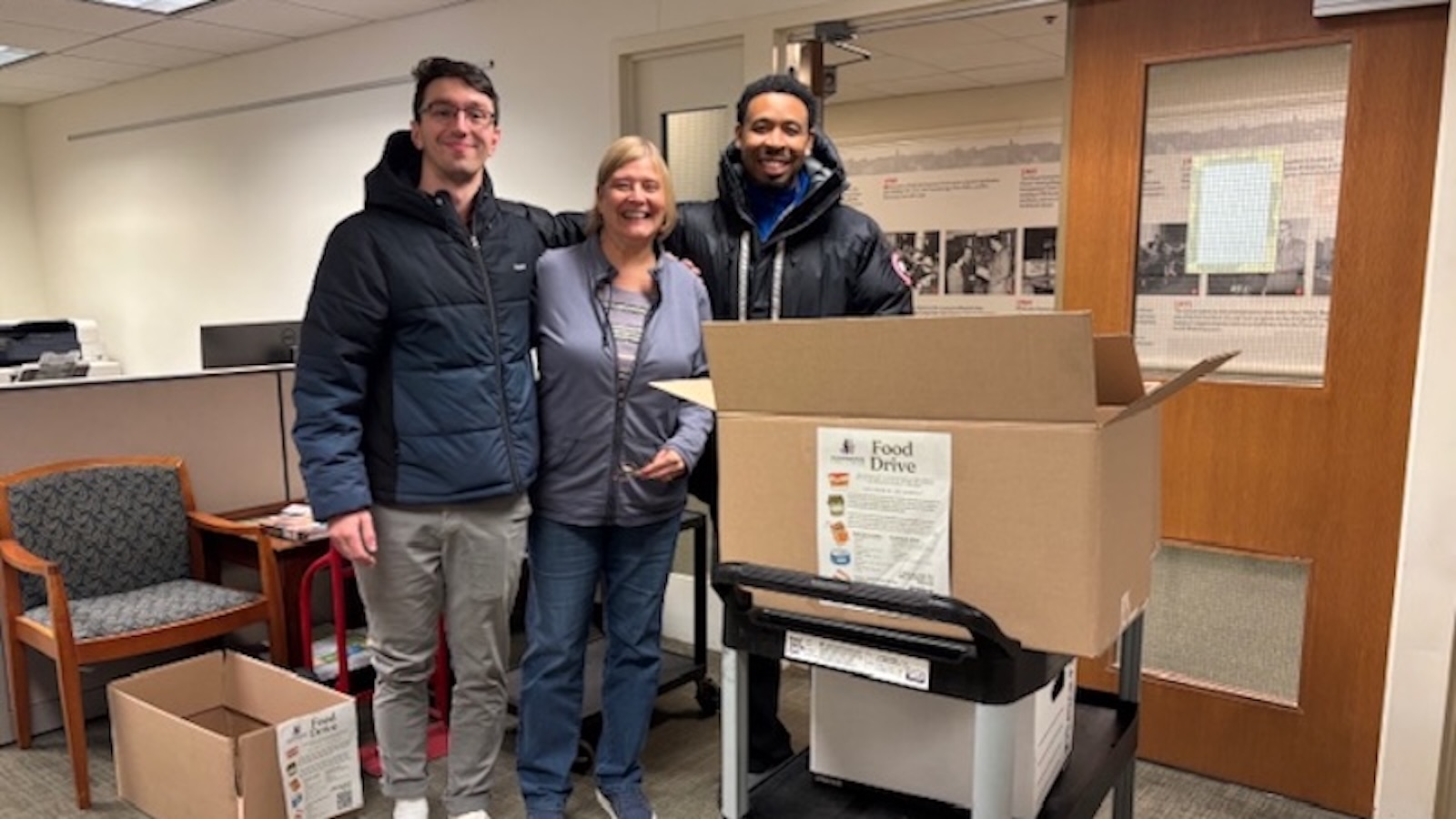 Three individuals standing indoors near a food drive collection area with large cardboard boxes labeled “Food Drive.”