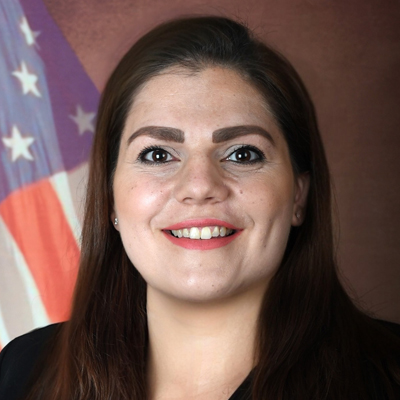 Professional headshot of a smiling woman in business attire with an American flag in the background.