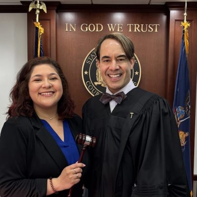 Two individuals standing in a courtroom setting. One is wearing a judicial robe and bow tie, while the other holds a gavel.