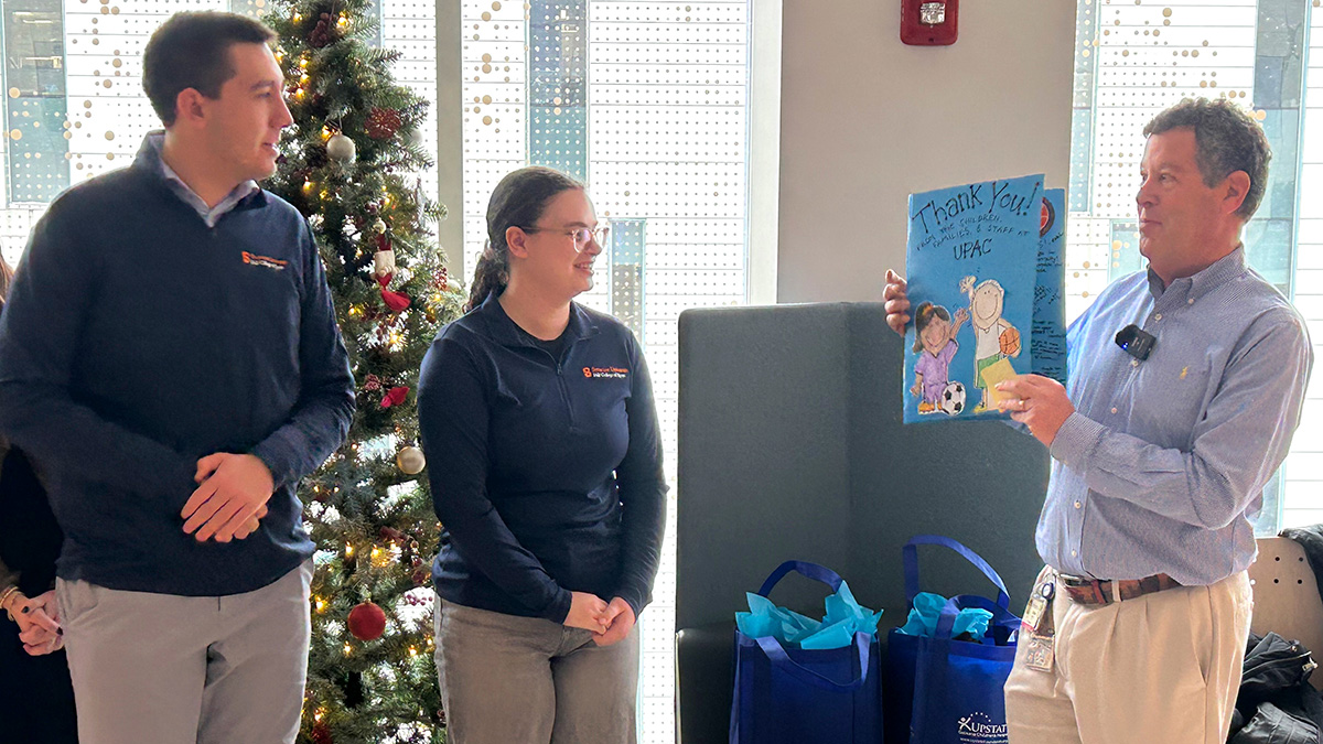 Three people stand near a decorated Christmas tree indoors while one person holds up a colorful thank-you card. Gift bags with blue tissue paper are visible on the floor.