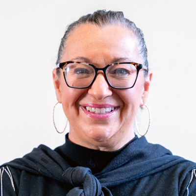 Professional headshot of a smiling woman with glasses and hoop earrings wearing a black turtleneck and dark blazer against a light background.