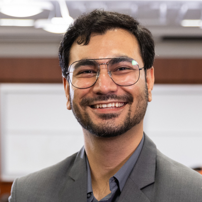 Professional headshot of a smiling man with glasses and a beard wearing a gray blazer and dark shirt in an office setting.