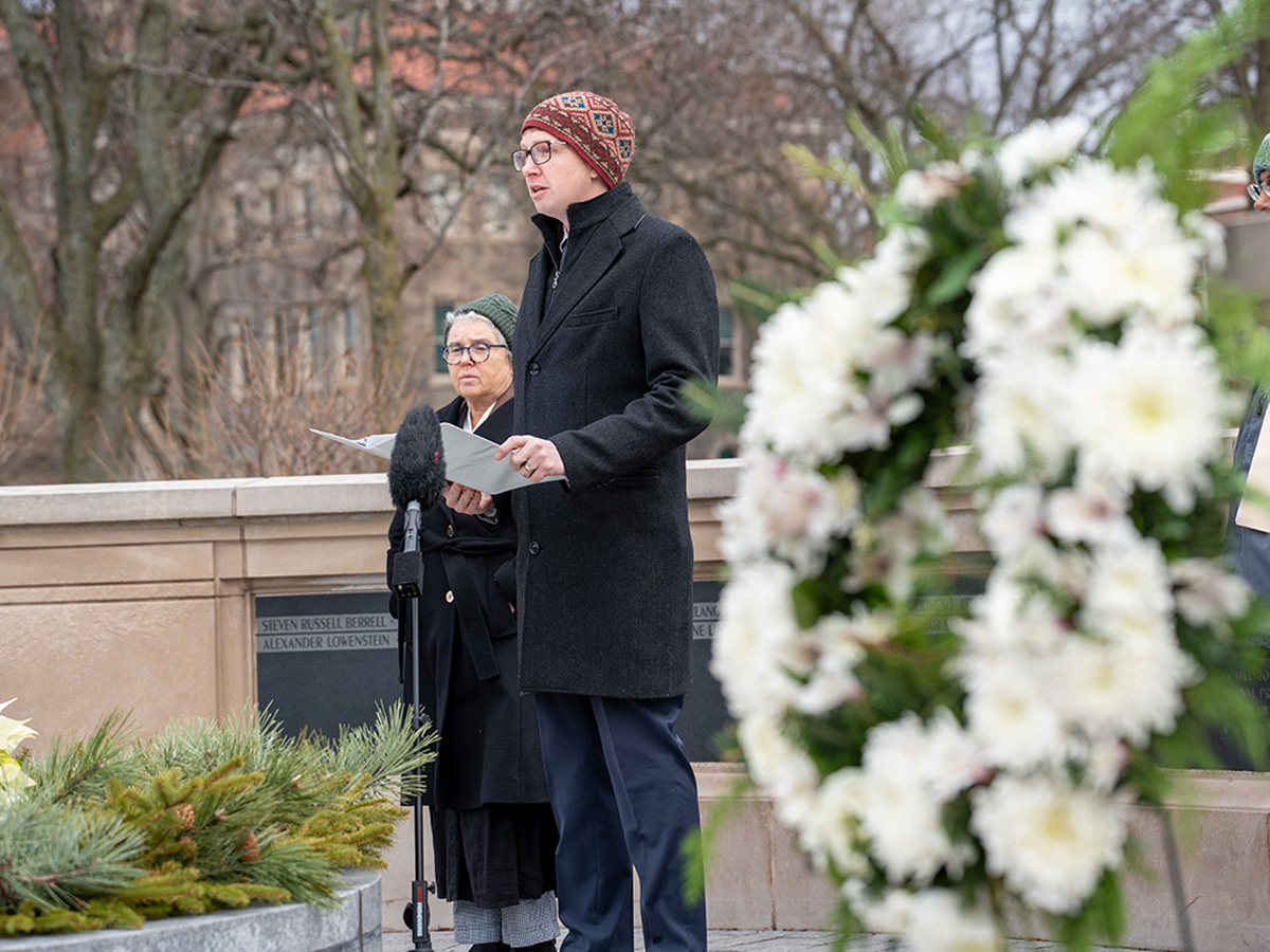 A man and a woman, bundled in warm coats, stand at the Wall of Remembrance. A floral wreath is off to the right