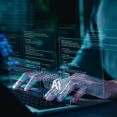 Hands typing on a laptop keyboard with multiple translucent holographic displays floating above, showing programming code, data visualizations, and an AI brain logo