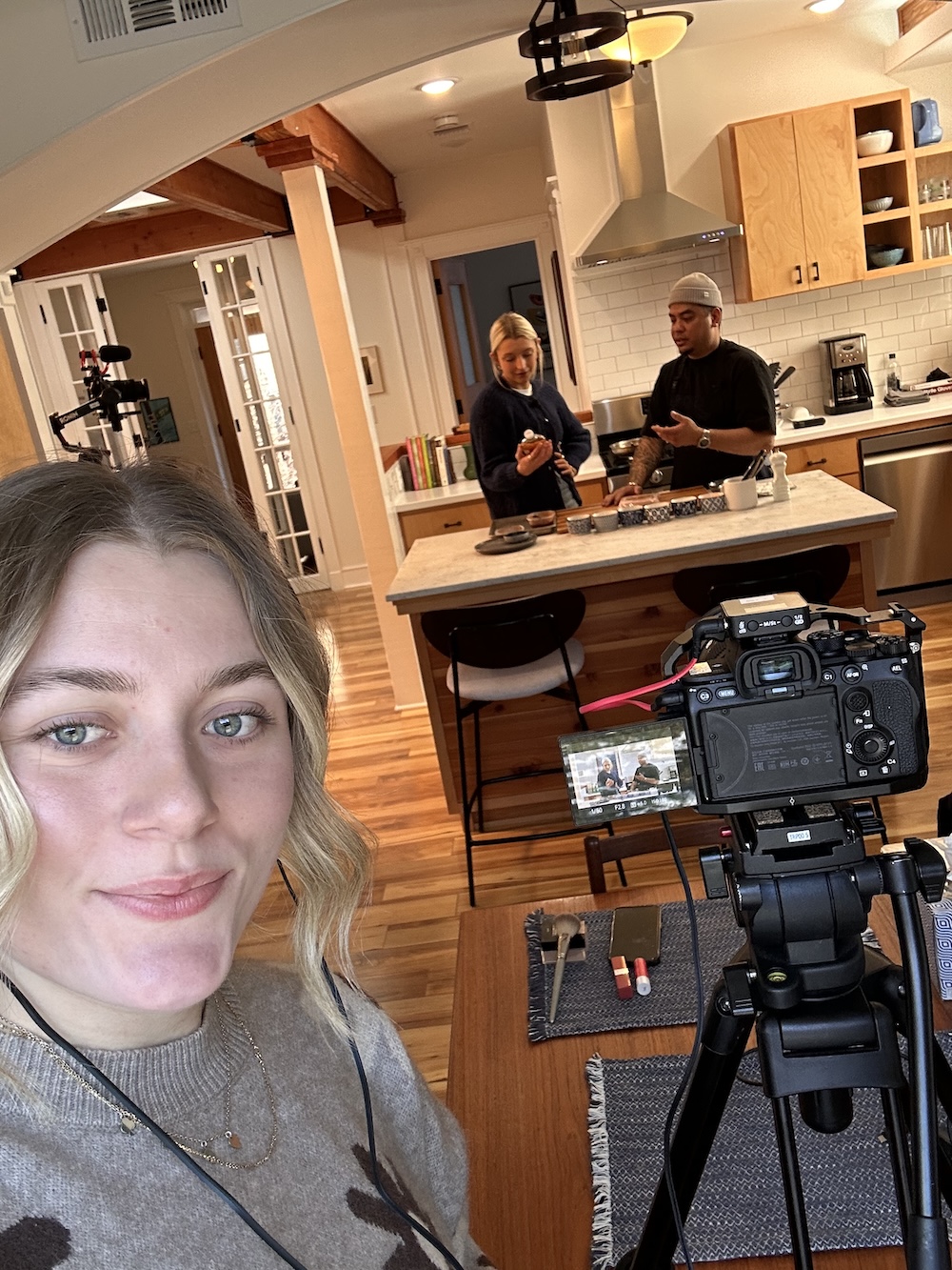 A behind-the-scenes view of a kitchen cooking demonstration being filmed, with cameras set up as two people prepare food at a kitchen island.