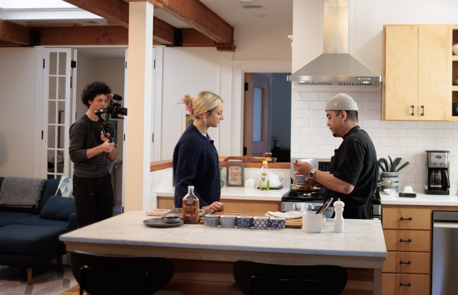 A home kitchen scene where a cook prepares food at the stove while another person stands nearby, and a videographer films the process 