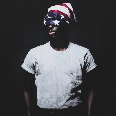 Portrait of a person wearing an American flag head wrap and white t-shirt against a dark background.