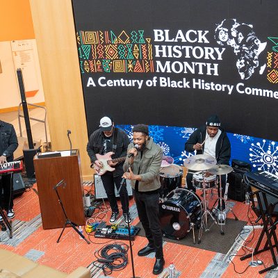 Musicians perform live on keyboards, guitar, and drums during a Black History Month celebration in an indoor event space.