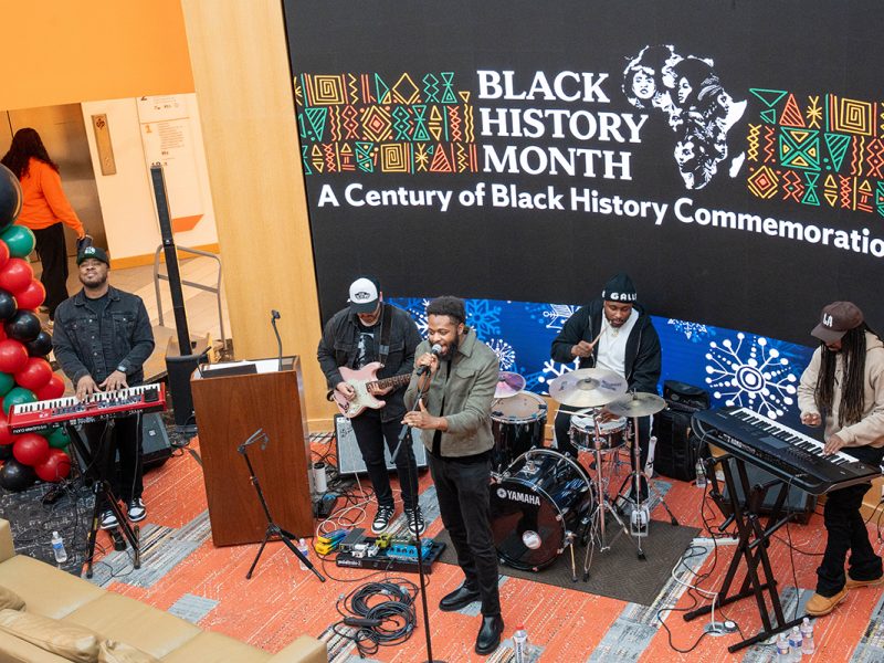 Musicians perform live on keyboards, guitar, and drums during a Black History Month celebration in an indoor event space.