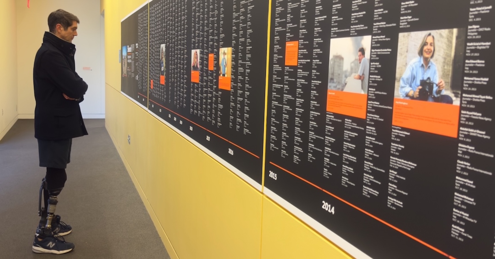 Person with prosthetic legs standing and looking at the memorial wall displaying names and a highlighted photo section.
