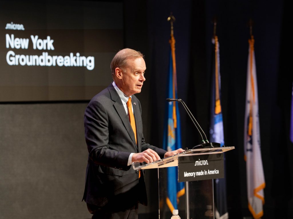 A person speaks at a podium during the Micron New York groundbreaking event, with flags displayed in the background.