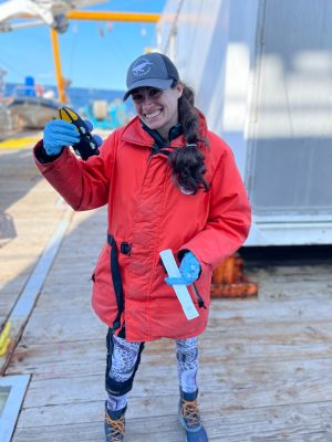 Marine researcher in red jacket and blue gloves holds a scientific instrument aboard a research vessel.