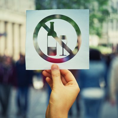 Hand holding a cut-out sign with a “no alcohol” symbol against a blurred city street background.