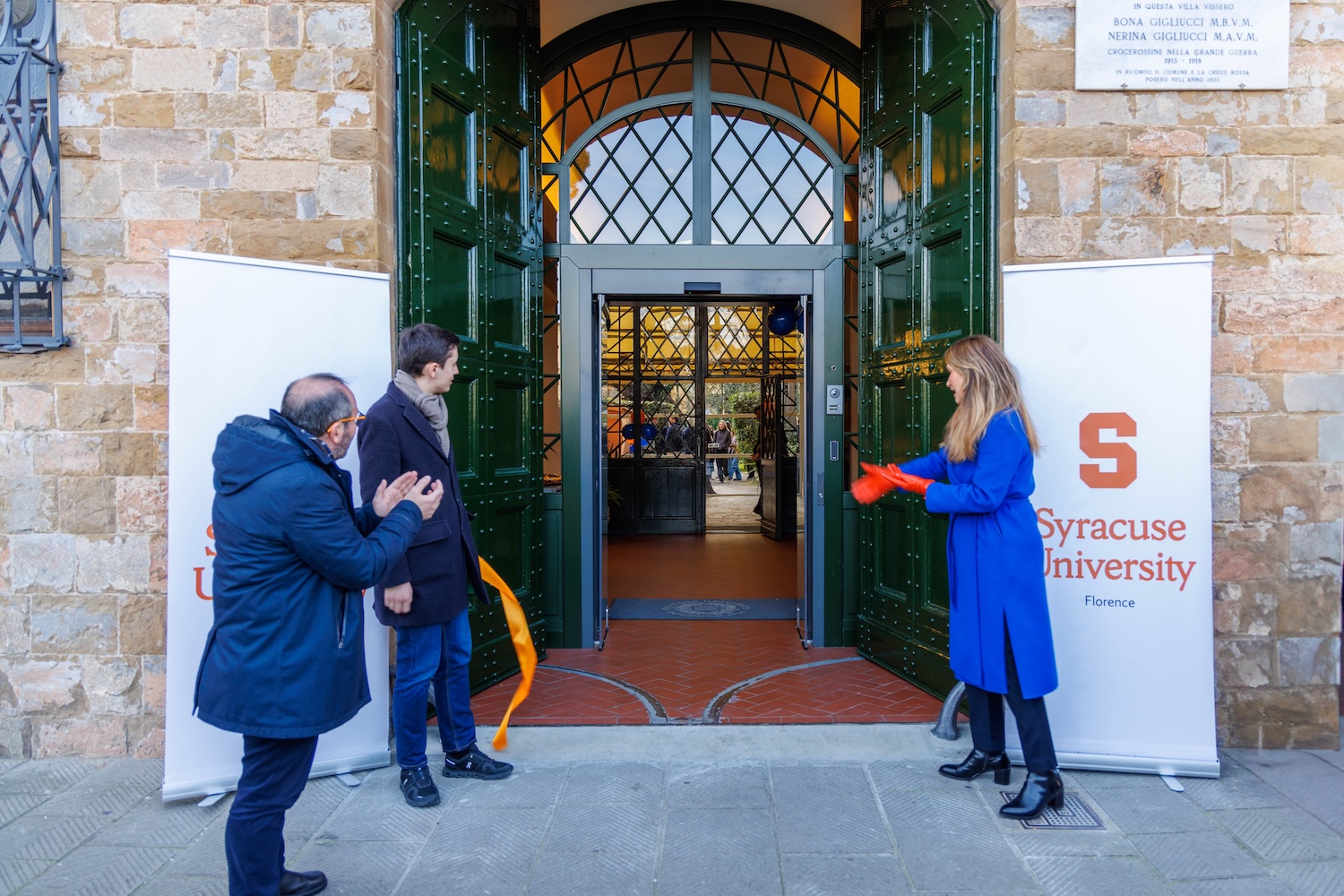 Three people stand at the entrance of a building between two Syracuse University Florence banners, holding a cut orange ribbon.